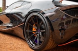 A close-up view of a shiny black sports car reveals intricate details of its wheel and tire. The metallic surface reflects nearby surroundings, including a photographer taking pictures. The vehicle is parked on a gravel surface, adding texture to the image.