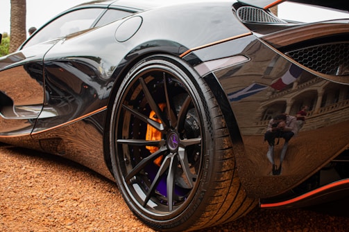 A close-up view of a shiny black sports car reveals intricate details of its wheel and tire. The metallic surface reflects nearby surroundings, including a photographer taking pictures. The vehicle is parked on a gravel surface, adding texture to the image.