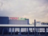 A large building with a sign reading 'TESCO Lotus' in colorful letters. The building features a sleek, modern design with a light gray facade. Below the sign is a pedestrian walkway where silhouettes of people are visible. The background shows a cloudy sky and some distant buildings.