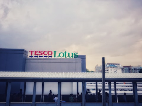 A large building with a sign reading 'TESCO Lotus' in colorful letters. The building features a sleek, modern design with a light gray facade. Below the sign is a pedestrian walkway where silhouettes of people are visible. The background shows a cloudy sky and some distant buildings.