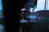 A person is standing on a sidewalk holding a purple umbrella. A car is passing by in the foreground. The traffic light is green, indicating go. A billboard with contact numbers is partially visible in the background.