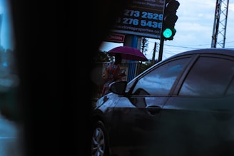 A person is standing on a sidewalk holding a purple umbrella. A car is passing by in the foreground. The traffic light is green, indicating go. A billboard with contact numbers is partially visible in the background.