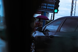 A person is standing on a sidewalk holding a purple umbrella. A car is passing by in the foreground. The traffic light is green, indicating go. A billboard with contact numbers is partially visible in the background.