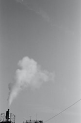 A black and white image featuring a factory smokestack emitting white smoke against a clear sky. The industrial structure includes a metal railing and an antenna, with a visible cable running diagonally across the frame.
