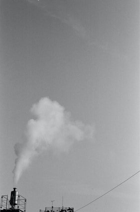 A black and white image featuring a factory smokestack emitting white smoke against a clear sky. The industrial structure includes a metal railing and an antenna, with a visible cable running diagonally across the frame.