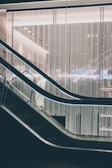 A modern escalator with sleek design lines moves through a contemporary building interior. The backdrop features a wall of vertical light strips, creating a futuristic and minimalistic aesthetic. The lighting is soft and ambient, highlighting the metallic and glass elements of the structure.