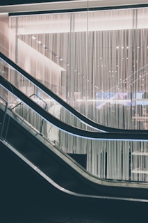 A modern escalator with sleek design lines moves through a contemporary building interior. The backdrop features a wall of vertical light strips, creating a futuristic and minimalistic aesthetic. The lighting is soft and ambient, highlighting the metallic and glass elements of the structure.