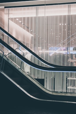 A modern escalator with sleek design lines moves through a contemporary building interior. The backdrop features a wall of vertical light strips, creating a futuristic and minimalistic aesthetic. The lighting is soft and ambient, highlighting the metallic and glass elements of the structure.