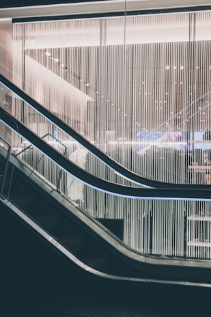 A modern escalator with sleek design lines moves through a contemporary building interior. The backdrop features a wall of vertical light strips, creating a futuristic and minimalistic aesthetic. The lighting is soft and ambient, highlighting the metallic and glass elements of the structure.