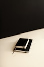 Two black notebooks with white pages are stacked on top of each other on a flat surface. The background consists of a black area transitioning into a beige or light tan area, creating a minimalistic and clean look.