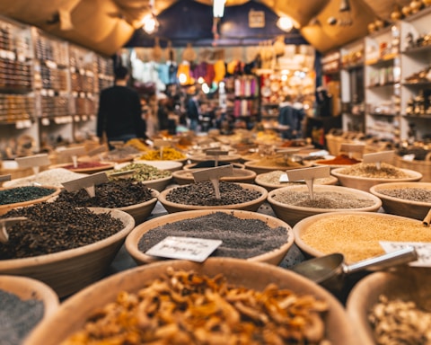 A vibrant market scene with numerous bowls filled with diverse spices and herbs, neatly arranged in rows. The market is bustling with activity, with a person blurred in the background walking past various other stalls filled with goods. Warm lighting creates an inviting and cozy atmosphere.