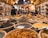 A vibrant market scene with numerous bowls filled with diverse spices and herbs, neatly arranged in rows. The market is bustling with activity, with a person blurred in the background walking past various other stalls filled with goods. Warm lighting creates an inviting and cozy atmosphere.