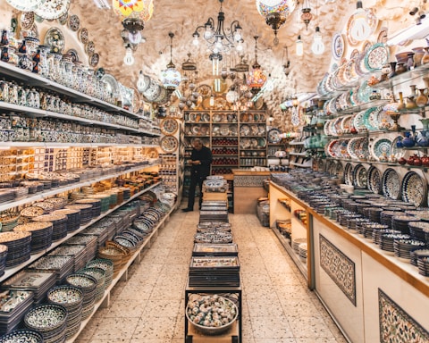 A vibrant, well-lit shop filled with a variety of intricately designed ceramic plates, bowls, and vases. The shelves are lined with colorful pottery featuring detailed patterns. Hanging lamps with stained glass add a warm ambiance. A person can be seen standing among the pottery, contributing to the bustling and inviting atmosphere.