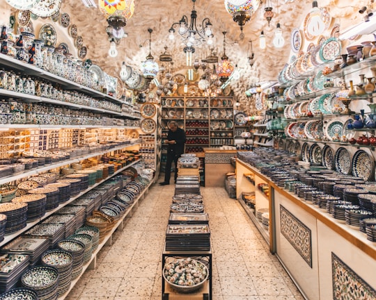 A vibrant, well-lit shop filled with a variety of intricately designed ceramic plates, bowls, and vases. The shelves are lined with colorful pottery featuring detailed patterns. Hanging lamps with stained glass add a warm ambiance. A person can be seen standing among the pottery, contributing to the bustling and inviting atmosphere.