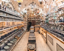 A vibrant, well-lit shop filled with a variety of intricately designed ceramic plates, bowls, and vases. The shelves are lined with colorful pottery featuring detailed patterns. Hanging lamps with stained glass add a warm ambiance. A person can be seen standing among the pottery, contributing to the bustling and inviting atmosphere.