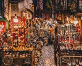 A bustling market scene filled with a variety of intricate metal crafts, jewelry, and textiles. Warm lighting from hanging bulbs illuminates the vibrant colors and detailed patterns of the fabrics draped around the shop. Shelves are crowded with ornate lamps, small sculptures, and an array of traditional ornaments. The narrow aisle adds to the feeling of abundance and richness in the display.
