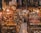 A bustling market scene filled with a variety of intricate metal crafts, jewelry, and textiles. Warm lighting from hanging bulbs illuminates the vibrant colors and detailed patterns of the fabrics draped around the shop. Shelves are crowded with ornate lamps, small sculptures, and an array of traditional ornaments. The narrow aisle adds to the feeling of abundance and richness in the display.