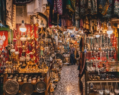 A bustling market scene filled with a variety of intricate metal crafts, jewelry, and textiles. Warm lighting from hanging bulbs illuminates the vibrant colors and detailed patterns of the fabrics draped around the shop. Shelves are crowded with ornate lamps, small sculptures, and an array of traditional ornaments. The narrow aisle adds to the feeling of abundance and richness in the display.