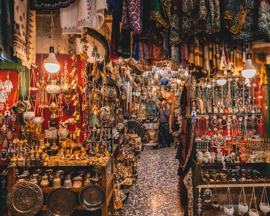 A bustling market scene filled with a variety of intricate metal crafts, jewelry, and textiles. Warm lighting from hanging bulbs illuminates the vibrant colors and detailed patterns of the fabrics draped around the shop. Shelves are crowded with ornate lamps, small sculptures, and an array of traditional ornaments. The narrow aisle adds to the feeling of abundance and richness in the display.