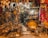 A densely packed antique shop filled with an eclectic mix of brass and bronze objects. The shelves are lined with various artifacts, including menorahs, plates, pitchers, and vases. Vibrant fabrics hang from above, adding a splash of color. The space appears narrow and cluttered with items stacked and hung closely together.