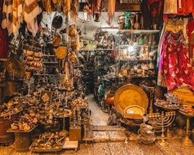 A densely packed antique shop filled with an eclectic mix of brass and bronze objects. The shelves are lined with various artifacts, including menorahs, plates, pitchers, and vases. Vibrant fabrics hang from above, adding a splash of color. The space appears narrow and cluttered with items stacked and hung closely together.