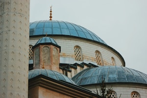 Detail of a mosque's architecture featuring multiple blue domes and a tall minaret. The structure has ornate designs and decorative elements like crescent finials.