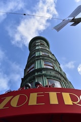 A cylindrical building with a green patina finish towers against a clear blue sky. The building features multiple bay windows and is topped by a red curved canopy with the word 'ZOETRO' prominently displayed in yellow letters. Overhead, a utility wire and a sign can be seen, and fluffy clouds accentuate the bright sky.
