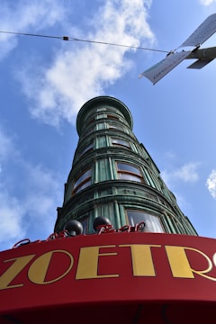 A cylindrical building with a green patina finish towers against a clear blue sky. The building features multiple bay windows and is topped by a red curved canopy with the word 'ZOETRO' prominently displayed in yellow letters. Overhead, a utility wire and a sign can be seen, and fluffy clouds accentuate the bright sky.