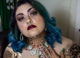 A person with vibrant blue hair and detailed facial makeup features prominently. They have facial piercings, including a septum ring and a nose ring, as well as bold, dark lipstick. The person is wearing ornate jewelry, including large earrings and a necklace. Tattoos are visible on their neck and shoulder, with intricate designs. Their nails are painted green.