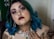 A person with vibrant blue hair and detailed facial makeup features prominently. They have facial piercings, including a septum ring and a nose ring, as well as bold, dark lipstick. The person is wearing ornate jewelry, including large earrings and a necklace. Tattoos are visible on their neck and shoulder, with intricate designs. Their nails are painted green.