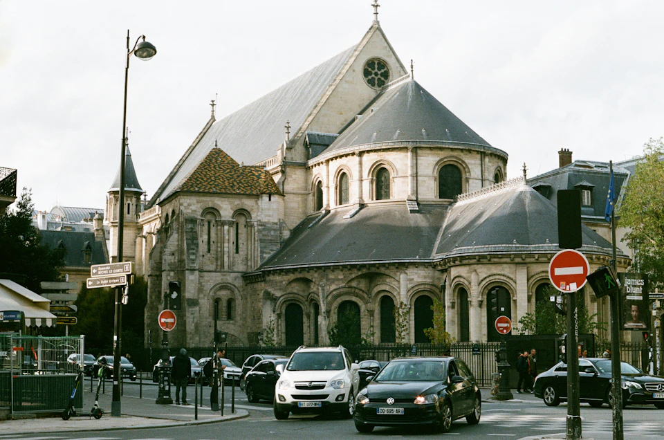 The image depicts a large, Gothic-style church with a tall steeple and a pointed roof, situated on a street corner in Paris, France. The church's architectural style is evident from its intricate details and the use of traditional materials such as stone and wood. Several cars are parked along the street, including a white SUV and a black sedan, adding to the urban atmosphere. Pedestrians can be seen walking on the sidewalk, contributing to the lively city scene.