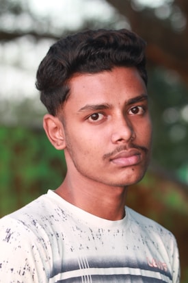 A young man with short, styled hair and a mustache is looking directly at the camera. He is wearing a light-colored shirt with a minimalist pattern, and the background appears to be a blend of natural green and brown, likely outdoors.