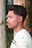 A young man with short, styled hair is in profile view. He is wearing a light-colored patterned shirt and is standing outdoors in front of a natural background with tree trunks and greenery.