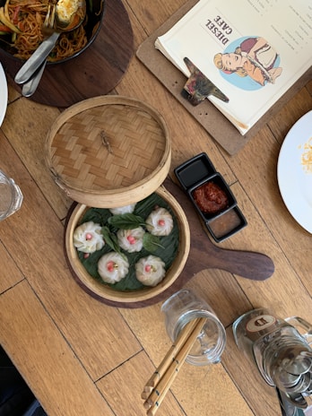 A wooden table with a bamboo steamer basket containing six dumplings garnished with green leaves and small pieces of red garnish. Next to it is a small dish with sauce divided into two sections: a thick red sauce and a dark liquid sauce. There is also a menu on a clipboard with retro artwork, a white plate with some food remnants, and an empty glass mug with chopsticks inside.