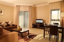 A well-furnished living room features a brown sofa with two white cushions, a dark wooden coffee table with a fruit arrangement, and a matching area rug. A television is on a wooden stand against the wall. To the right, there's a dining table set for three with cups and saucers. The room is softly lit through beige curtains covering large windows, creating a warm and inviting atmosphere.