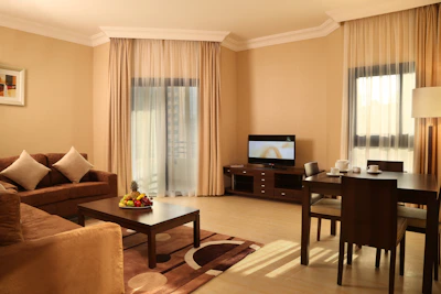 A well-furnished living room features a brown sofa with two white cushions, a dark wooden coffee table with a fruit arrangement, and a matching area rug. A television is on a wooden stand against the wall. To the right, there's a dining table set for three with cups and saucers. The room is softly lit through beige curtains covering large windows, creating a warm and inviting atmosphere.