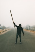 A person stands in the middle of an empty road, holding a long stick or pole in one hand. The individual is wearing a hooded jacket and a mask, possibly to protect against dust or pollution. The environment appears hazy and overcast, suggesting smog or mist, with sparse vegetation and structures visible in the background.