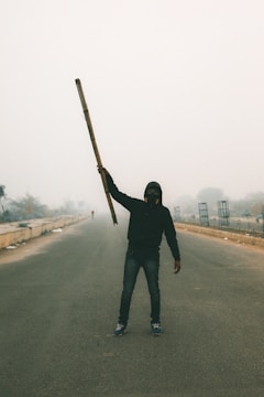 A person stands in the middle of an empty road, holding a long stick or pole in one hand. The individual is wearing a hooded jacket and a mask, possibly to protect against dust or pollution. The environment appears hazy and overcast, suggesting smog or mist, with sparse vegetation and structures visible in the background.