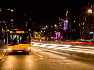 A city street at night with a yellow city bus parked on the side, displaying the route number 111 with the destination 'Gocław'. Bright, blurred lights from passing vehicles create light streaks on the road. Skyscrapers and buildings with illuminated signage are visible in the background, including a tower with purple lighting.