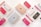 A flat lay arrangement of various Apple products including iPhones with different colored cases, iPads, and Apple Watch bands spread across a light background. The devices and accessories are neatly organized, showcasing a palette of pink, red, and neutral tones.