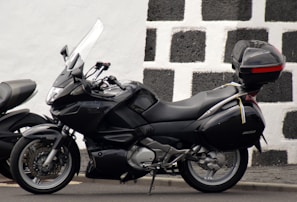 A sleek black motorcycle is parked against a contrasting checkered wall that is made of black and white stone. The motorcycle features a windshield, side mirrors, and a storage box mounted on the rear end. The overall design suggests a touring model with comfortable seating and aerodynamic features.