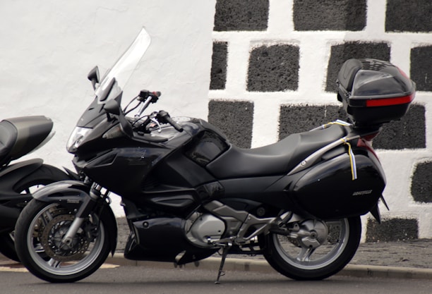 A sleek black motorcycle is parked against a contrasting checkered wall that is made of black and white stone. The motorcycle features a windshield, side mirrors, and a storage box mounted on the rear end. The overall design suggests a touring model with comfortable seating and aerodynamic features.