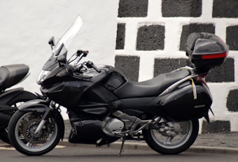 A sleek black motorcycle is parked against a contrasting checkered wall that is made of black and white stone. The motorcycle features a windshield, side mirrors, and a storage box mounted on the rear end. The overall design suggests a touring model with comfortable seating and aerodynamic features.