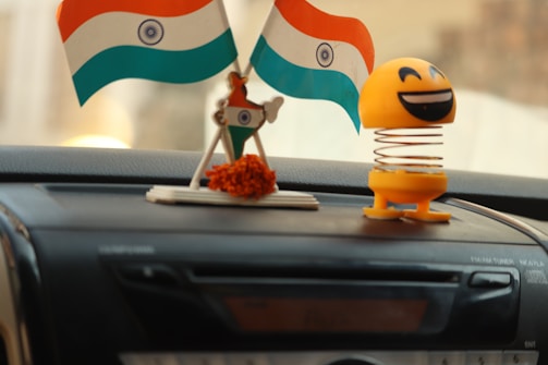 Two small Indian flags are displayed on a car dashboard next to a smiling yellow spring-mounted toy. The flags are attached to a decorative base with orange flowers. The dashboard has a CD player or stereo system situated directly beneath the objects.