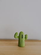 A small, green cactus-shaped object sits on a light wooden surface against a plain gray background. The object has a slot, suggesting it might be a decorative piggy bank.