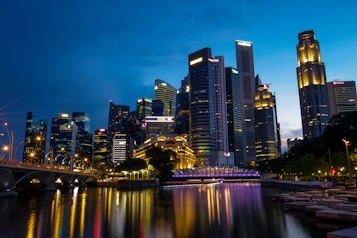 A vibrant cityscape featuring a skyline of illuminated skyscrapers reflecting on a calm waterfront. The scene transitions from day to night with a deep blue sky, creating a serene yet lively atmosphere. Streetlights and building lights add warmth against the cool tones of the evening.