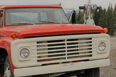 A vintage red Ford truck is parked with a slightly rusty front and white grill. It has a classic round headlight design, and the background features a forest with tall pine trees.
