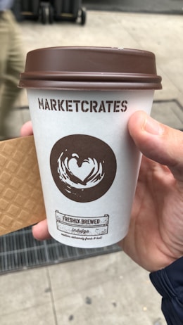 A hand holds a takeaway coffee cup with a brown lid and white sleeve. The cup features a stylized heart logo and the text 'MARKETCRATES' and 'FRESHLY BREWED.' A waffle-textured sleeve partially covers the side. The background shows a city sidewalk and a blurred figure walking past.