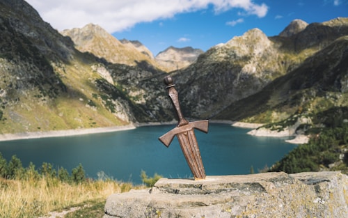 A sword embedded in a stone with a scenic backdrop of mountains and a tranquil lake under a clear blue sky.
