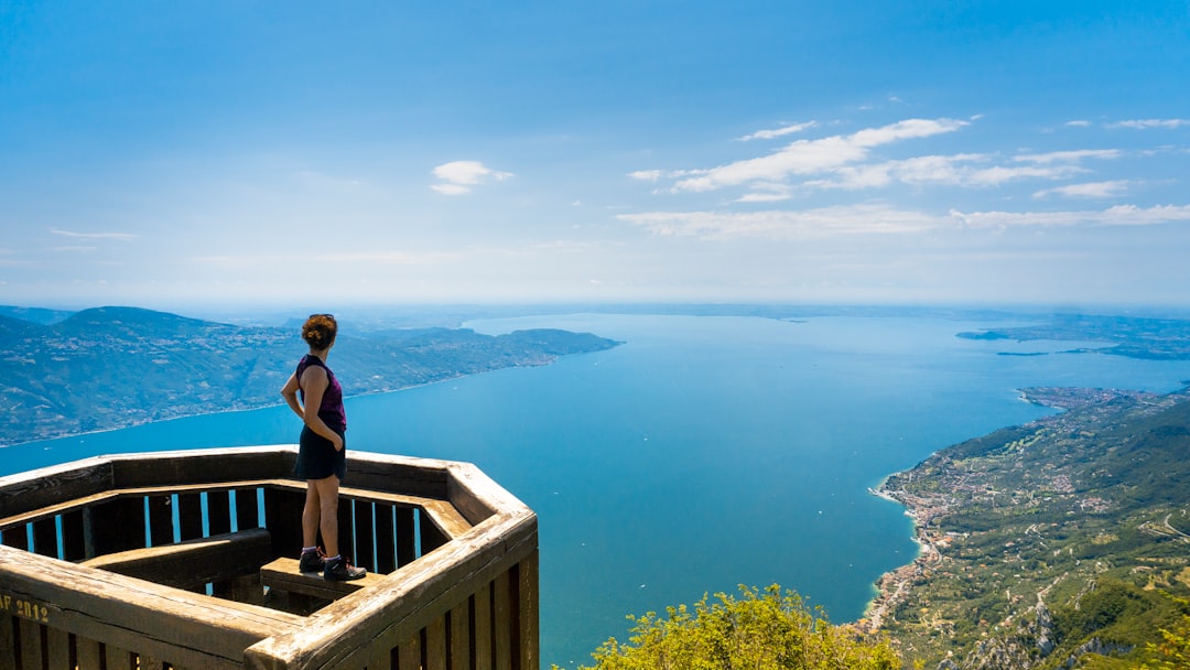 woman wearing black tank top standing near railings viewing blue sea and mountain under blue and white sky during daytime